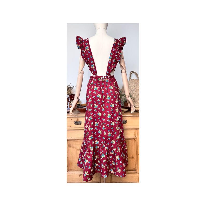 May include: A burgundy-colored dress with a floral pattern, featuring ruffled shoulder straps and a flared skirt. The dress is displayed on a mannequin, showcasing its vintage style. The dress has a waist measurement of approximately 28 inches.