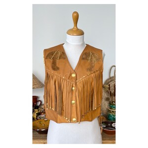 May include: A brown leather waistcoat with fringe and gold accents. The waistcoat has a pointed collar and a button closure. The waistcoat is decorated with two brown leather appliques on the front.