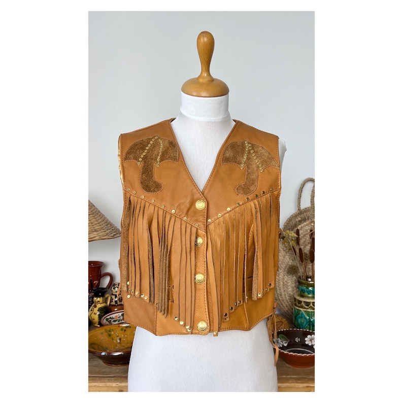 May include: A brown leather waistcoat with fringe and gold accents. The waistcoat has a pointed collar and a button closure. The waistcoat is decorated with two brown leather appliques on the front.