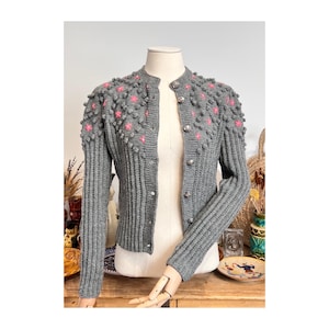 Könnte beinhalten: Graue Strickjacke im Vintage-Stil mit rundem Kragen und langen Ärmeln. Der Pullover hat rosa Blumenakzente und dekorative silberne Knöpfe. Die Ärmel und Schultern haben ein strukturiertes, erhabenes Design.