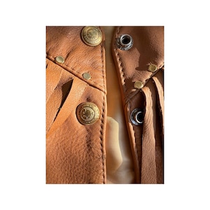 May include: Close-up of a brown leather waistcoat with fringe detailing. The waistcoat has multiple gold-toned metal accents, including buttons and studs.