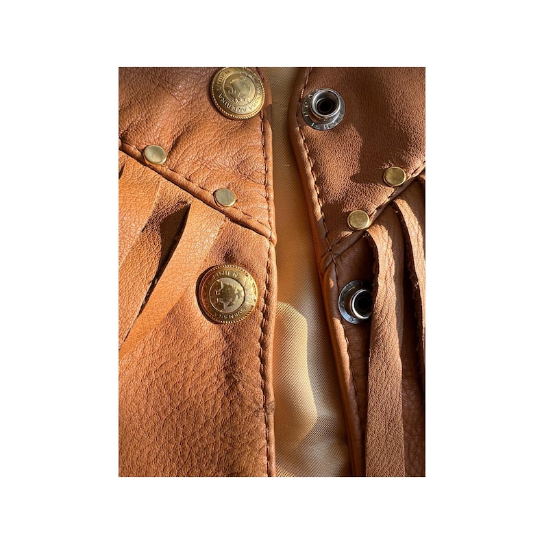 May include: Close-up of a brown leather waistcoat with fringe detailing. The waistcoat has multiple gold-toned metal accents, including buttons and studs.
