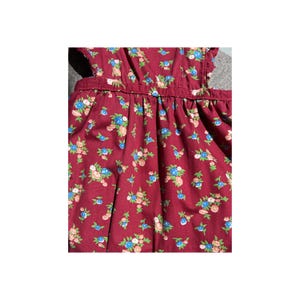 May include: A close-up of a burgundy dress with a floral pattern. The dress features small blue, pink, and white flowers with green leaves. The fabric is gathered at the waist and has a ruffled edge.