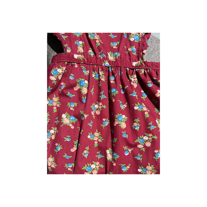May include: A close-up of a burgundy dress with a floral pattern. The dress features small blue, pink, and white flowers with green leaves. The fabric is gathered at the waist and has a ruffled edge.