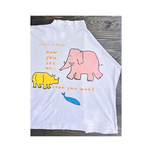 Op de afbeelding: Wit T-shirt met een hoge halslijn met een roze olifant, gele neushoorn en blauwe walvis illustratie. De tekst "animals in danger" en "now you see us... soon you won't" is gedrukt in oranje en geel.