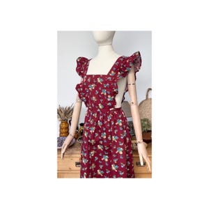 May include: A burgundy floral print dress with ruffled sleeves and straps. The dress features a square neckline and a fitted waist. The fabric is patterned with small blue and white flowers. The dress is displayed on a mannequin.
