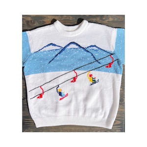 Pode incluir: Camisola de malha branca com um design de estação de esqui. A camisola apresenta um design de blocos de cores azul e branco com esquiadores bordados, teleféricos e paisagem de montanha. As mangas são azul claro.