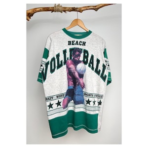 Könnte beinhalten: Ein grau-grünes T-Shirt mit einem Grafikdruck eines Mannes, der Beachvolleyball spielt. Der Text "Beach Volleyball" ist in Grün auf das Shirt gedruckt. Der Text "Crazy Wave Sports System" ist in Schwarz auf das Shirt gedruckt.