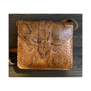 Peut inclure: Un sac à bandoulière de forme carrée fabriqué en cuir de serpent marron avec un motif texturé et écailleux. Le sac présente un rabat frontal avec un détail de sangle verticale centrale et une fine bandoulière assortie fixée sur les côtés.