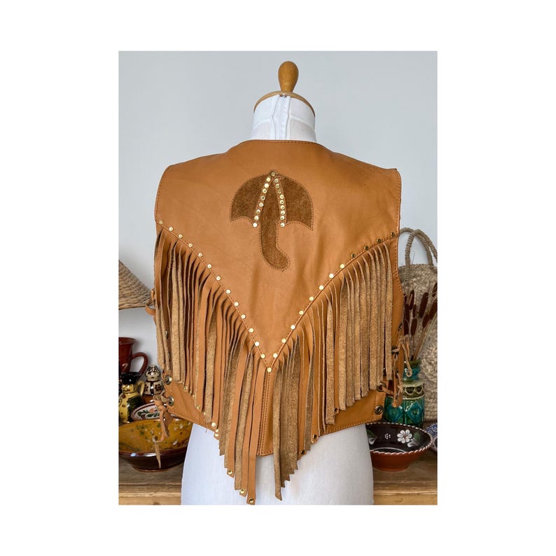 May include: A brown leather vest with fringe and a suede umbrella design on the back. The vest is decorated with gold studs.