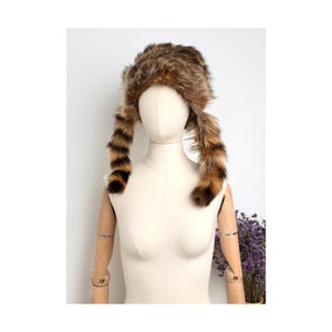 Può includere: Un cappello in pelliccia sintetica marrone e nero con corona arrotondata e due lunghi pannelli laterali pendenti con motivo a righe. Il cappello è esposto su una testa e un busto di manichino di colore neutro, che mettono in risalto la pelliccia.