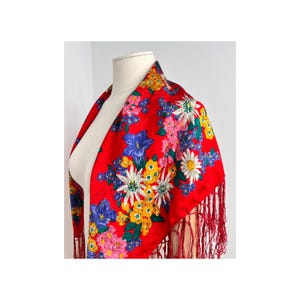 May include: A red floral scarf with a vibrant pattern of blue, white, yellow, and pink flowers. The scarf has a fringed edge and is displayed on a mannequin. The scarf is a square shape.