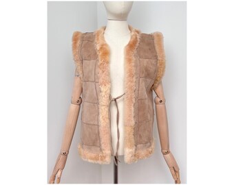 May include: A tan sheepskin vest with a light brown suede exterior and a cream shearling lining. The vest has a square-stitched design and a tie closure. The edges are trimmed with fluffy shearling.