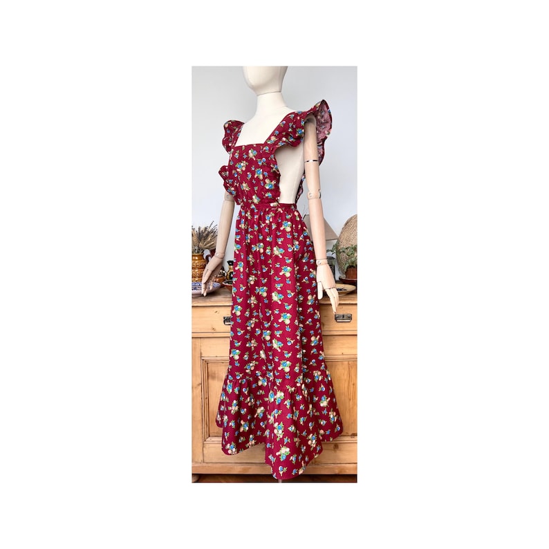 May include: A burgundy floral print dress with ruffled shoulder straps and a tiered skirt. The dress features a square neckline and a fitted bodice, displayed on a mannequin. The dress is a vintage style.