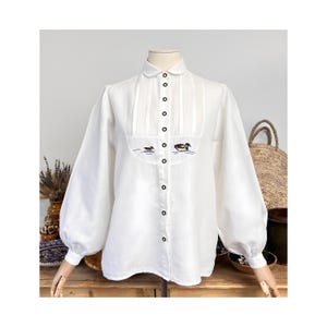 May include: White blouse with a round collar and long, puffy sleeves. The front features pleats and an embroidered design of two ducks on a pond. The blouse has black buttons down the front and on the cuffs.