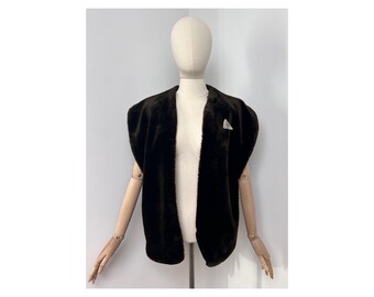 May include: A dark brown faux fur vest with a shawl collar and a small, silver-toned triangular brooch. The vest is displayed on a mannequin against a plain white background. The vest has a sleeveless design.
