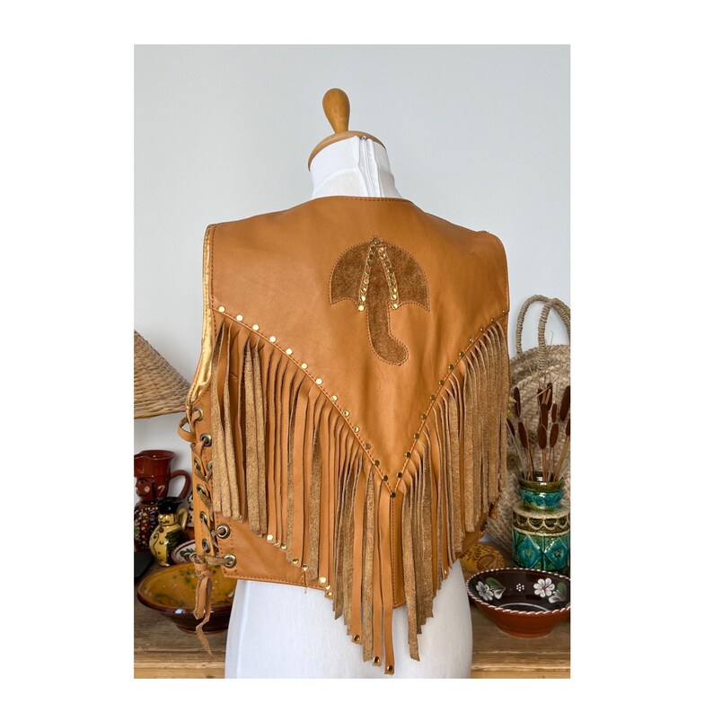 May include: A brown leather waistcoat with fringe and gold studs. The waistcoat has a suede patch on the back with a gold umbrella design.