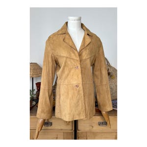 Puede incluir: Una chaqueta de ante marrón con cierre de botón y detalles de costura. La chaqueta está en un maniquí y tiene mangas largas.