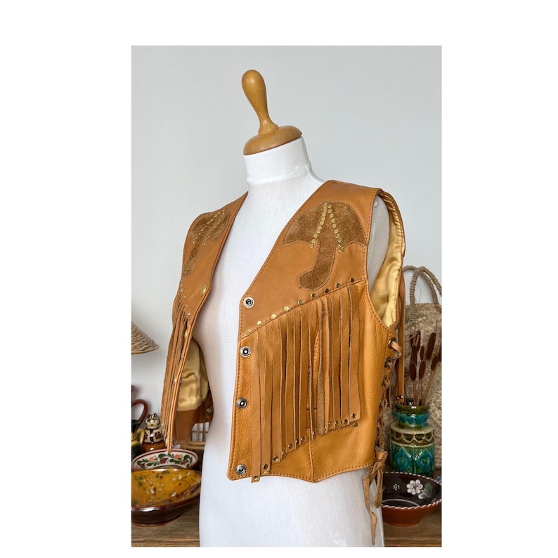 May include: A brown leather vest with fringe and gold studs. The vest has a suede patch on the front with a geometric design.