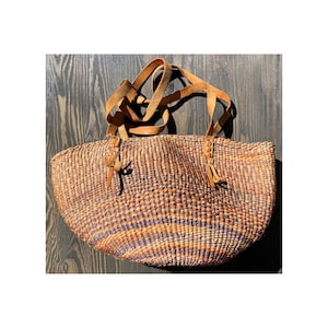 Può includere: Una borsa a cesta intrecciata di forma arrotondata, con una miscela di tonalità arancioni, marroni e viola. Ha due manici in pelle marrone chiaro e nappe decorative in pelle. La borsa è su una superficie di legno scuro.