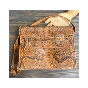 Peut inclure: Un sac à main vintage en peau de serpent avec une poignée tressée et une bandoulière. Le sac a une forme rectangulaire avec une fermeture à rabat et une poche avant. Le motif en peau de serpent est un mélange de marron, de beige et de noir.