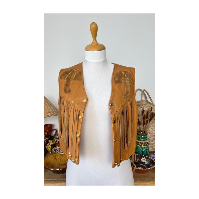 May include: A brown leather waistcoat with fringe and decorative stitching. The waistcoat has a pointed collar and a front zip closure.