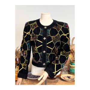 Puede incluir: Chaqueta de ante negra con un diseño de patchwork único. La chaqueta presenta formas geométricas en varios colores y texturas, delineadas con costuras amarillas. Tiene un cuello redondo y botones decorativos.