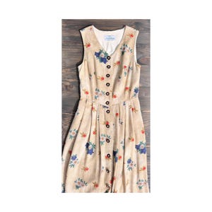 Könnte beinhalten: Beigefarbenes ärmelloses Kleid mit floralem Muster in Blau, Rot und Weiß. Das Kleid hat einen V-Ausschnitt, eine Knopfleiste und einen Faltenrock. Das Vintage-Kleid hat ein tailliertes Oberteil und ein weißes Futter.