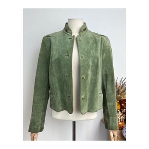 Op de afbeelding: Een groene suède jas met een opstaande kraag en knopen. De jas heeft lange mouwen en kleine zakken. De jas wordt op een mannequin getoond.