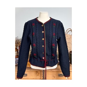 May include: A navy blue knit cardigan featuring vertical cable knit patterns, embroidered red flower motifs, and a round neckline. The garment has a front button closure with wooden buttons, a red drawstring waist tie, and green trim detailing.