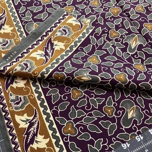 May include: A folded piece of fabric featuring a deep purple background with a repeating pattern of grey leaves and tan flowers. A wide border in tan and dark brown displays stylized floral motifs. A black ruler shows measurements in inches.