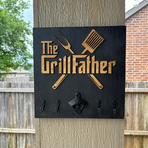 May include: Black wooden sign with the text "The Grill Father" in gold lettering. The sign features a crossed spatula and grill fork, and has four hooks and a bottle opener.
