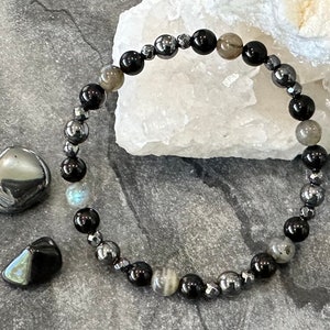 May include: A black and silver beaded bracelet with black, silver, and iridescent beads. The bracelet is on a gray surface with a white rock in the background.