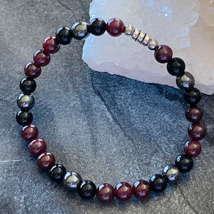 May include: A beaded bracelet with alternating red, black, and silver beads. The bracelet is made of natural stones and is designed to be worn on the wrist.