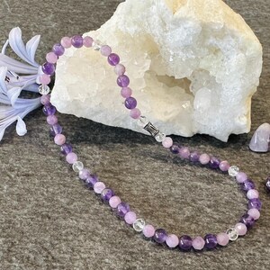 May include: A purple and pink amethyst gemstone necklace with a silver clasp. The necklace is on a gray surface with a white crystal rock and purple amethyst stones.