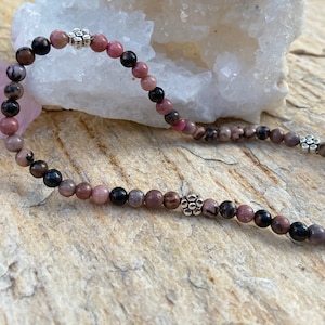 May include: A pink and black beaded bracelet with silver flower accents. The bracelet is made with natural stones and is approximately 7 inches in length.