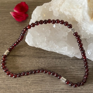 May include: A beaded necklace composed of small, round, deep red garnet stones. The necklace is strung together with a silver clasp featuring a decorative design. The necklace is displayed on a wooden surface with a white crystal formation in the background.