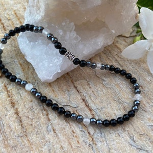 May include: A black and silver beaded bracelet with a silver charm. The bracelet is made with black onyx, hematite, and white moonstone beads.