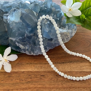 May include: A delicate white beaded necklace with a subtle iridescent sheen. The necklace is displayed on a wooden surface, with a large blue crystal and white flowers in the background. The necklace is a simple, elegant piece.