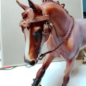 May include: A brown and white horse figurine with a leather bridle and leg wraps. The horse has a white stripe down its face and is posed with one leg raised. The bridle and leg wraps are brown.