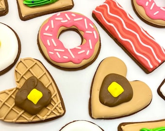 May include: Assortment of decorated cookies in various shapes and designs. Includes a pink frosted donut with sprinkles, a bacon strip, a heart-shaped pancake with butter, and a fried egg. Other cookies feature green frosting and colorful sprinkles.
