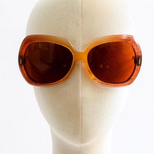 May include: Orange-tinted sunglasses with a retro, oversized frame. The lenses are a deep brown, and the frame has a gradient effect, transitioning from a lighter orange to a darker, almost tortoise-shell pattern. The sunglasses are displayed on a mannequin head.