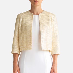 May include: A cream-coloured, cropped jacket with a textured, sequined surface. The jacket has three-quarter sleeves and a round neckline. The garment is worn over a white dress. The overall style is elegant and suitable for formal occasions.