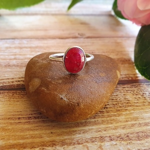 May include: A silver ring with a red oval gemstone set in the centre. The ring is resting on a brown rock.