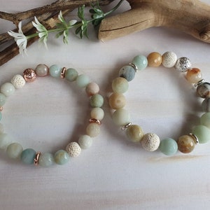 May include: Two bracelets made with green, brown, and white beads. One bracelet has a silver charm and the other has a rose gold charm.