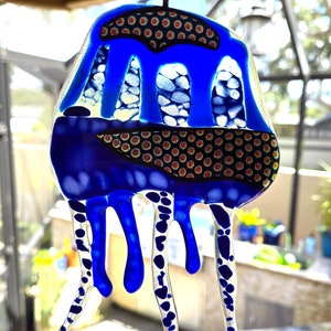 May include: A blue glass jellyfish with white and black polka dots. The jellyfish has a round body with a black and gold patterned band and long, thin tentacles.