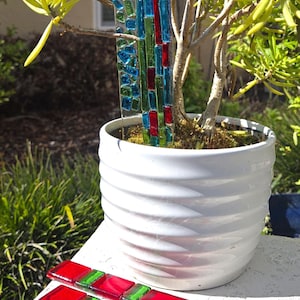 May include: A white ceramic pot with a plant and colorful glass garden stakes. The stakes feature red, green, and blue glass pieces. The plant has yellow-green leaves. Additional stakes are displayed in front of the pot.