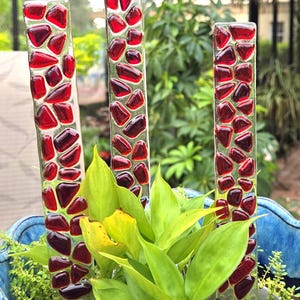 May include: Three glass garden stakes with red glass pieces set in clear glass. The stakes are placed in a blue ceramic planter with green plants. The stakes are approximately 12 inches tall.