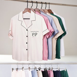 Op de afbeelding: Een set van acht korte mouwen knoophemden en acht paar shorts in verschillende kleuren, waaronder roze, blauw, wit, beige, groen en zwart. De shirts hebben een zwarte rand en een zak met de initialen "EP" geborduurd op de linker borst. De shorts hebben een zwarte rand rond de tailleband en de benen.