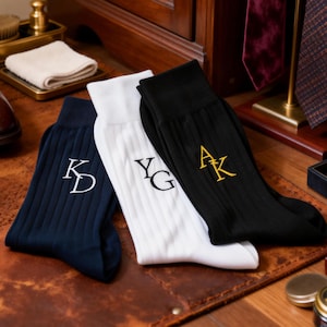 Puede incluir: Una colección de accesorios para hombre, que incluye zapatos de vestir de cuero marrón, tres pares de calcetines acanalados con iniciales bordadas y una selección de corbatas. Los calcetines son azul marino, blancos y negros. La escena está ambientada sobre una alfombra de cuero.